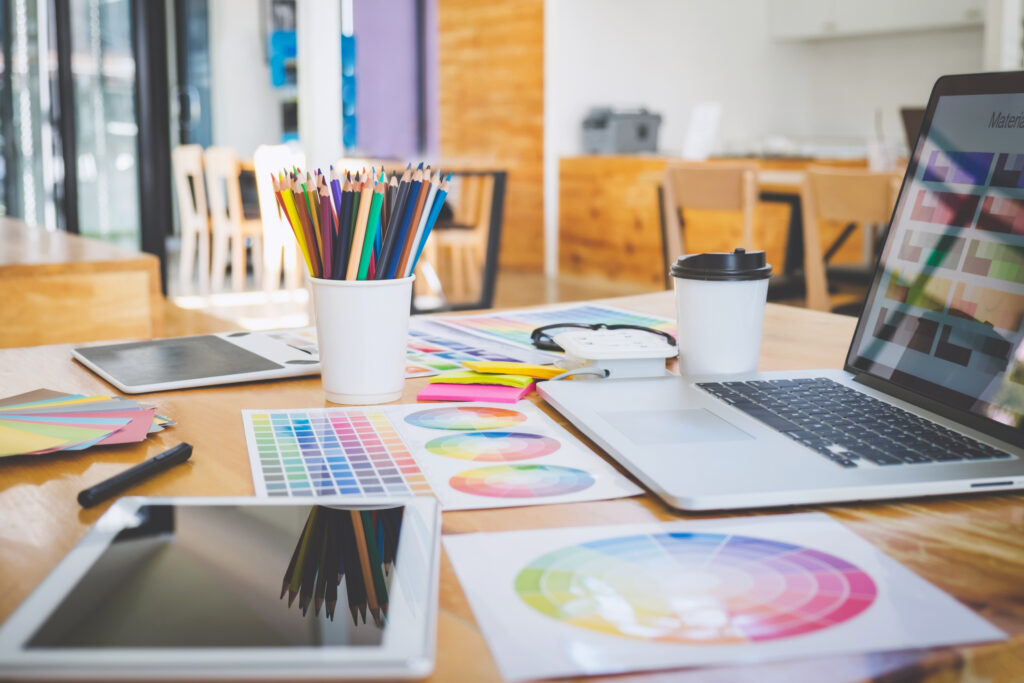 Color swatches, design tablets, a laptop, and colored pencils are spread across a graphic designer's desk.