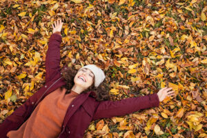 A woman wearing a customized jacket and beanie lies on autumn leaves with her arms outstretched.