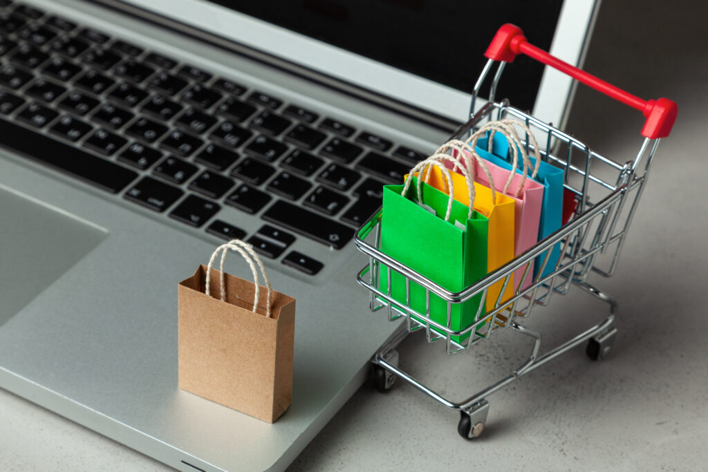 Online store concept: A miniature metal shopping cart full of paper bags is parked next to a laptop.