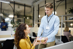 An employee hands his coworker a wrapped corporate gift at her workstation.